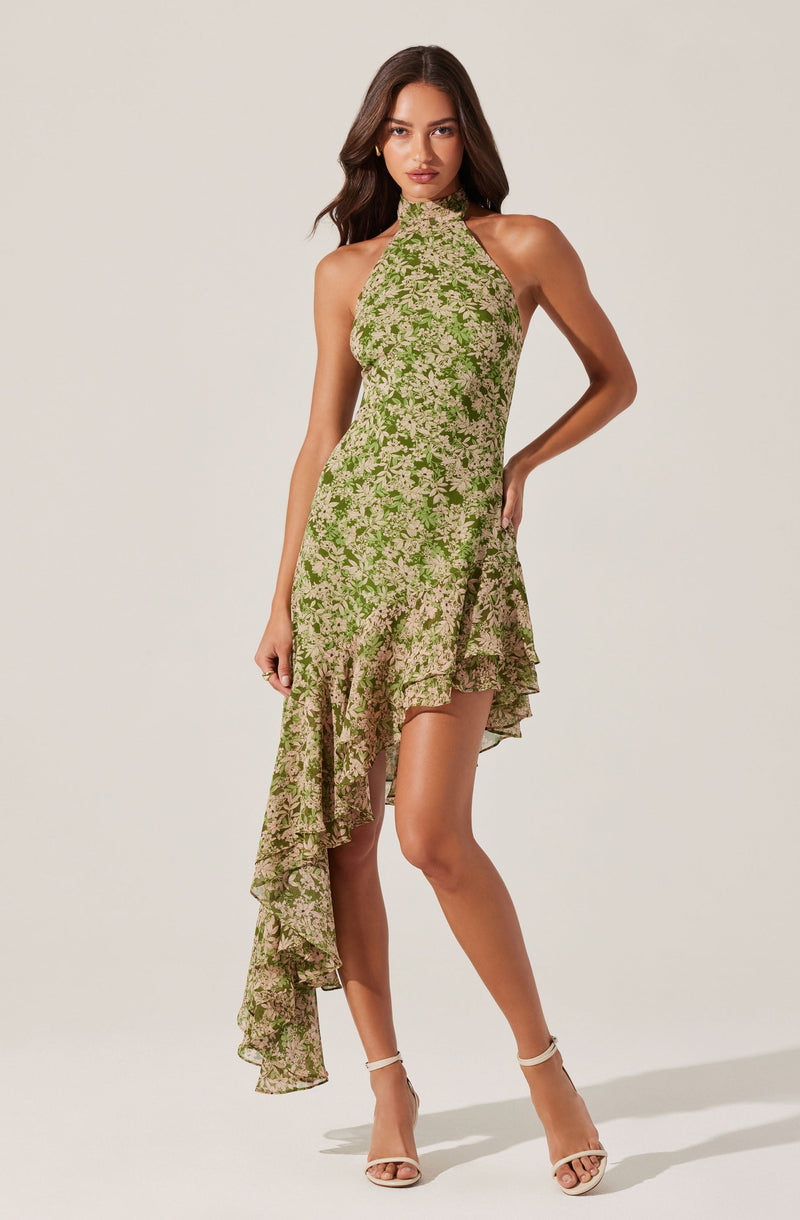 A woman is wearing a sleeveless, high-neck dress with an asymmetrical hemline. The dress features a green floral pattern with beige flowers, and has a ruffled detail along the lower hem on the left side. She is also wearing beige high-heeled sandals.