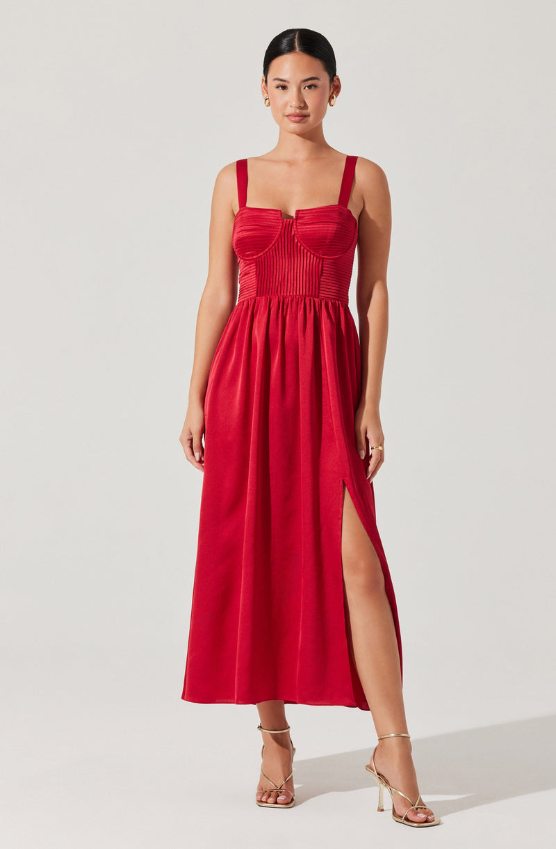 The dress is a sleeveless, floor-length gown in a bold red color. It features a fitted bodice with vertical pleats and structured, curved seam details that enhance the bust area. The dress has wide straps and a high slit on one side, revealing part of the leg. The skirt is gathered at the waist and flows down freely to the floor. The overall style is elegant and sophisticated.