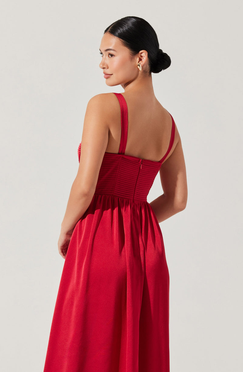 A woman is wearing a sleeveless, red dress with wide straps. The top of the dress features horizontal ruching and a fitted waist, with a zipper at the back. The skirt of the dress is gathered at the waist and flows down in a full, elegant style, made from a shiny, satin-like fabric. The woman has her hair styled in a neat bun and is wearing gold hoop earrings.