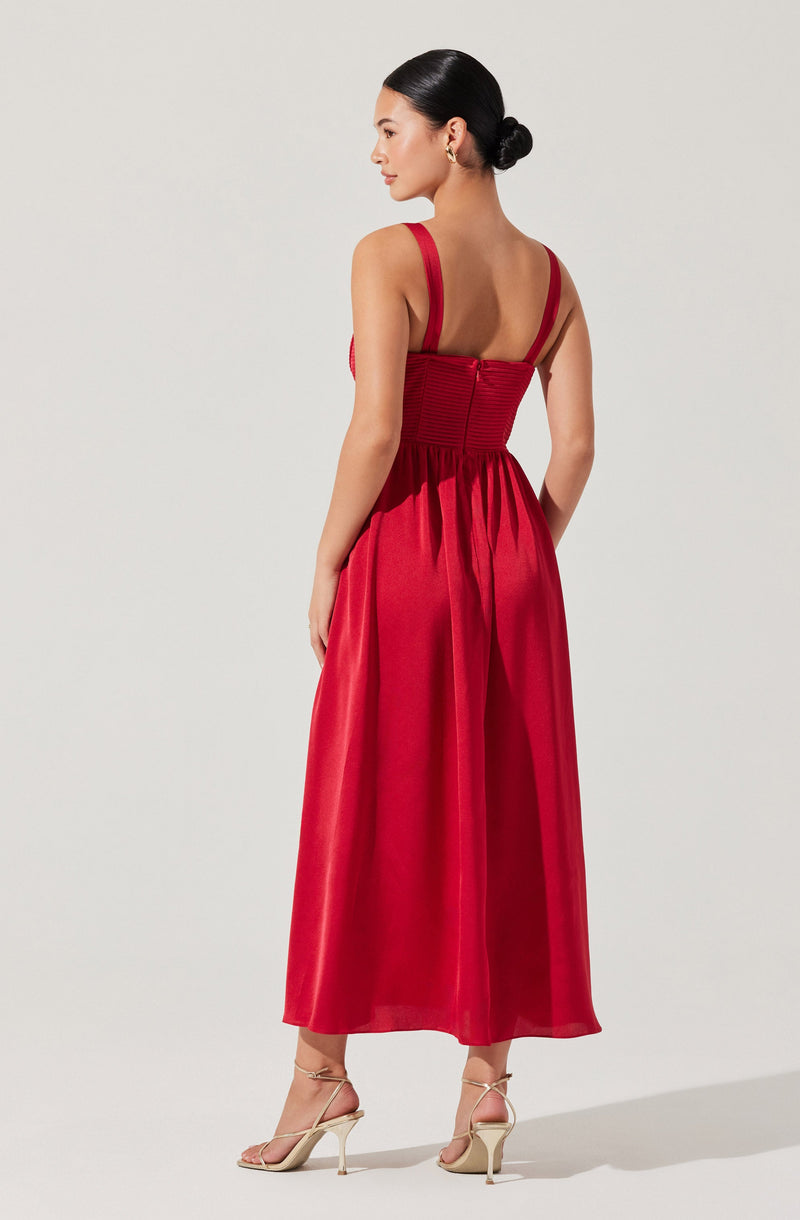 A woman is wearing a sleeveless, full-length red dress with wide shoulder straps. The bodice features horizontal pleats and a back zipper. The skirt is gathered at the waist, flowing down to the ankles. She is also wearing beige high-heeled sandals with ankle straps.