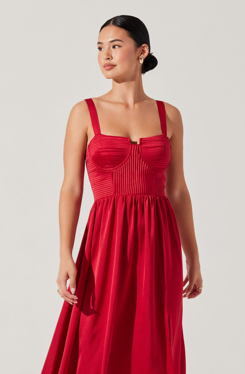 A woman is wearing a sleeveless, floor-length red dress with wide straps. The bodice of the dress features horizontal and vertical ribbed textures, creating a structured look. The skirt is gathered at the waist, flowing down in soft pleats.