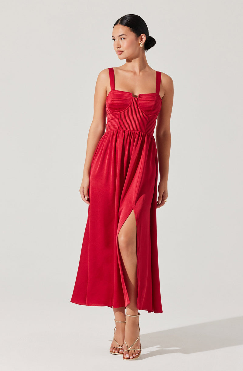A woman is wearing a long, sleeveless red dress with wide straps. The dress features a fitted bodice with vertical seam details, a centered hook-and-eye closure at the bust, and a high slit on one side that reveals her leg. She is also wearing beige high-heeled sandals.
