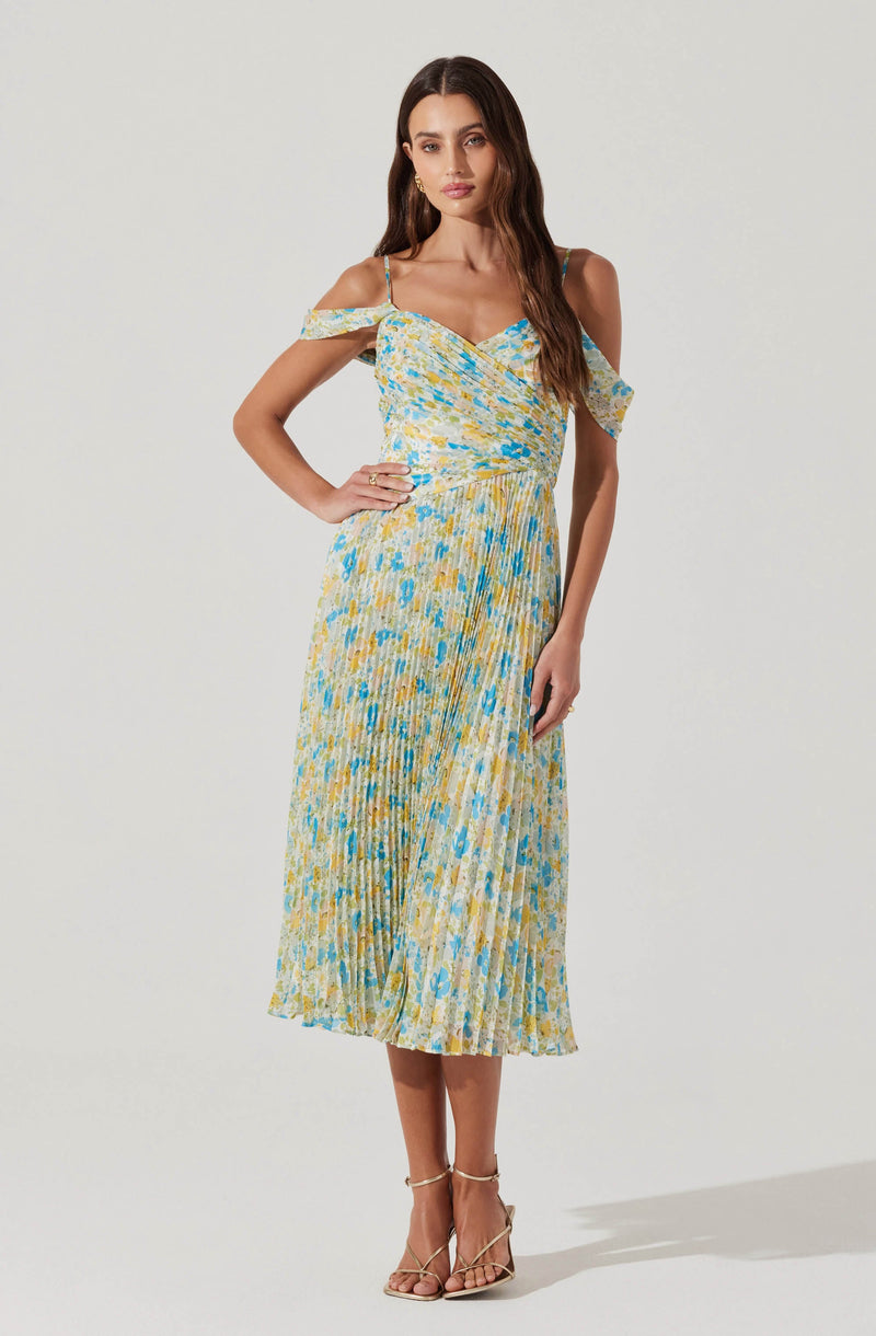 A woman is wearing a sleeveless, ankle-length dress with thin spaghetti straps and off-the-shoulder ruffle sleeves. The dress features a multicolored pattern in shades of yellow, blue, and white, with a fitted waist and a pleated skirt. She is also wearing strappy beige high-heeled sandals.