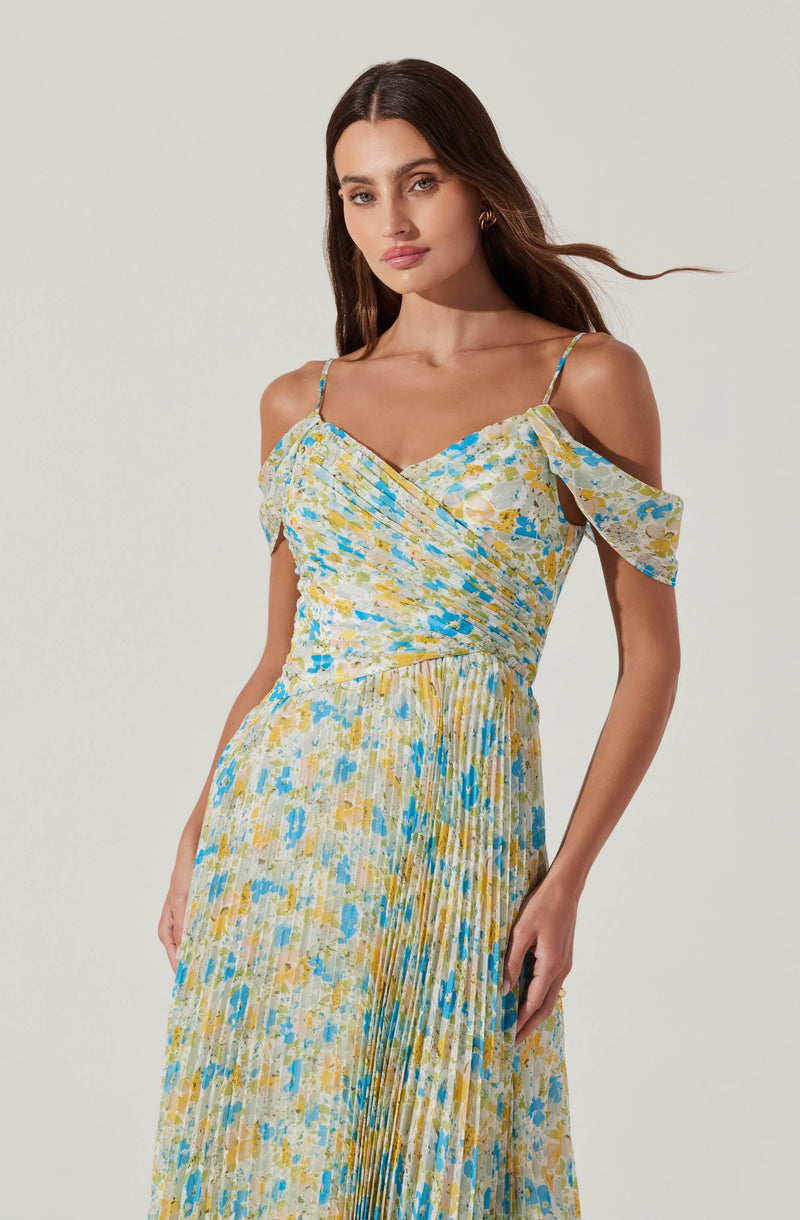 A woman is wearing a sleeveless, floral-printed dress with thin spaghetti straps. The dress has a ruffled, off-the-shoulder design on one side and features a multicolored pattern in shades of yellow, blue, and white. The fabric is lightweight and appears to have a pleated texture in the skirt portion.