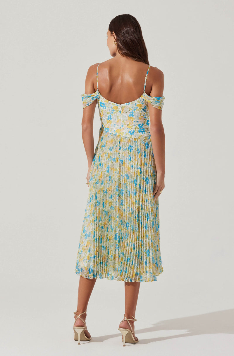 A woman is wearing a light-colored, floral print dress with shades of blue, yellow, and cream. The dress has thin spaghetti straps and off-the-shoulder details with small ruffled sleeves. The fabric appears lightweight and flowy, with a pleated skirt that reaches mid-calf.