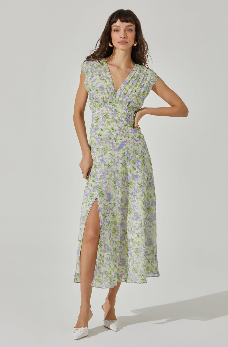 A woman is wearing a long, flowy dress with a floral pattern in shades of purple, green, and white. The dress has a V-neckline and short, slightly caped sleeves. It features a wrapped bodice and a high slit on the left side, revealing her leg. The fabric appears lightweight and semi-transparent. She is also wearing white shoes with a bow detail.