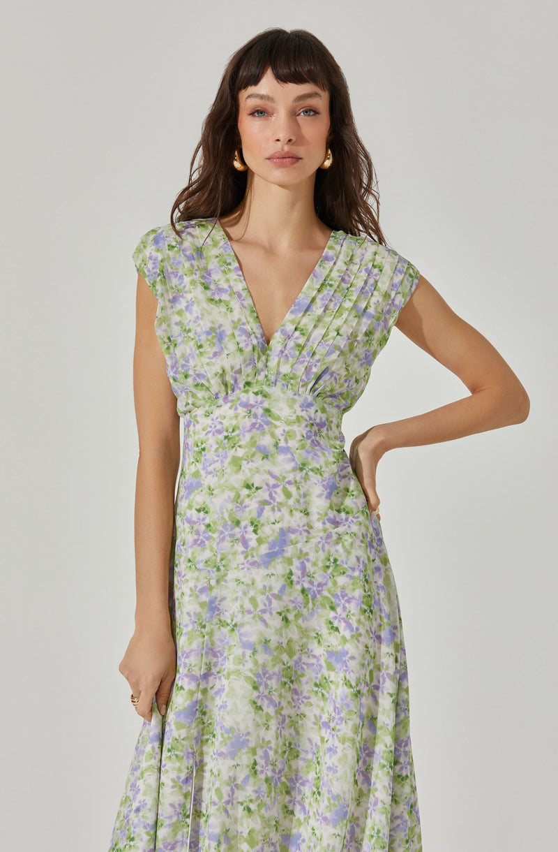 A woman is wearing a sleeveless, V-neck dress with a fitted bodice and a flowing skirt. The dress features a light pastel floral pattern in shades of purple, green, and white.