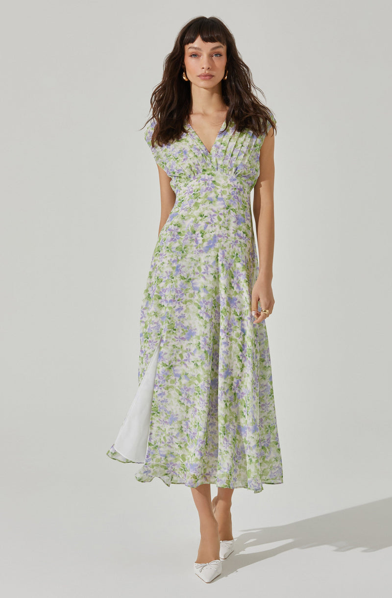 A woman is wearing a sleeveless, ankle-length dress with a V-neckline. The dress features a light purple and green floral pattern on a sheer fabric, with a white lining underneath. The dress has a front slit that reveals a white inner layer. She is also wearing white pointed-toe shoes with a bow detail.
