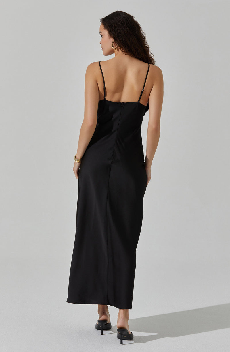 A woman is wearing a long, black satin dress with thin, adjustable spaghetti straps. The dress has a fitted bodice and falls straight to the floor. She is also wearing black heeled sandals.