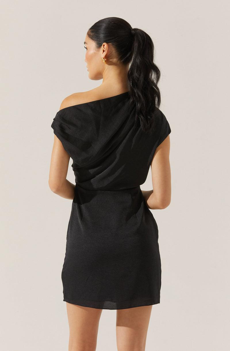 A woman is wearing a black satin dress with a one-shoulder design. The dress has a loose, draped sleeve on one side and is fitted at the waist, ending above the knees. She has dark hair styled in a high, wavy ponytail and is accessorized with small gold hoop earrings.