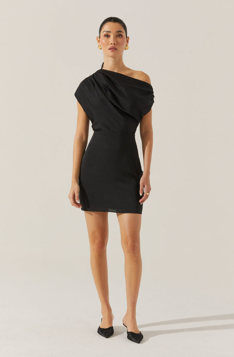 The woman is wearing a stylish black dress with an asymmetrical off-the-shoulder neckline. The dress has a draped, gathered detail on the upper part and a fitted, body-hugging silhouette that ends above the knees. She completes her look with black pointed-toe shoes featuring ruched detailing.