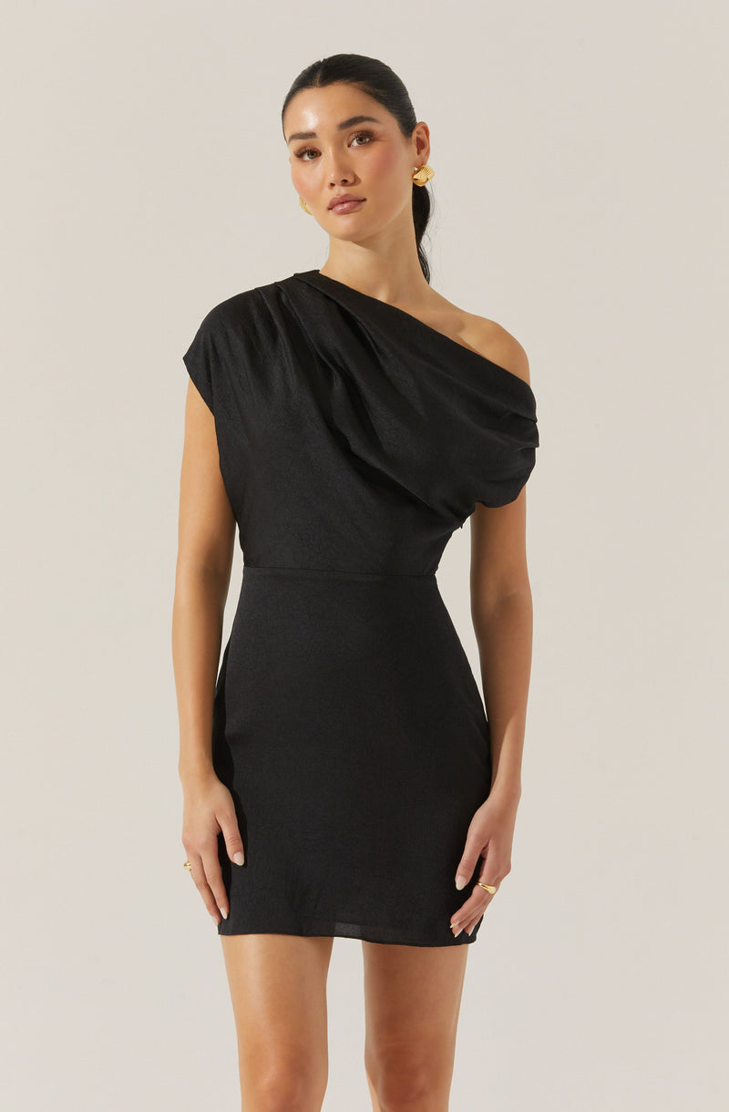 A woman is wearing a black dress with an asymmetrical design. The dress features a one-shoulder neckline with gathered fabric on the shoulder, creating draped detailing across the chest and shoulder area. The fitted dress is made of a sleek, slightly shiny fabric that accentuates her waist and hips.