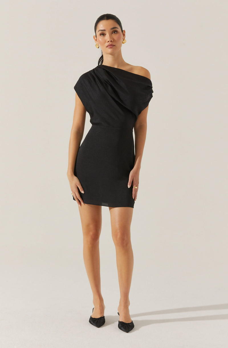A woman is wearing a black dress with an asymmetrical design. The dress has a draped, off-the-shoulder style on one side with a loose, gathered fabric over the shoulder, and fits snugly around the waist, ending above the knees. She is also wearing black pointed-toe shoes and gold hoop earrings.