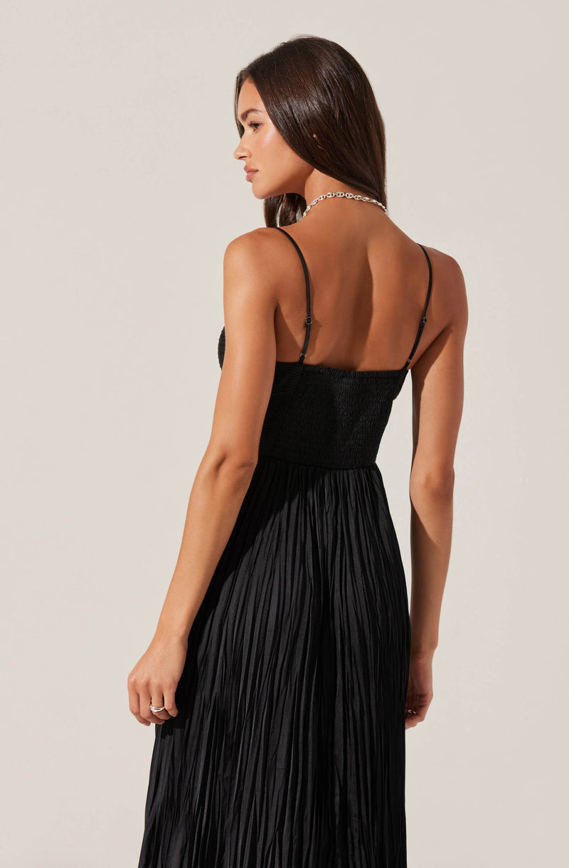 A woman is wearing a black spaghetti strap dress with a fitted bodice and a flowing, pleated skirt. The dress has thin straps over her shoulders, and she accessorizes with a silver chain necklace. Her long, dark brown hair is worn down, and she is posed with her back slightly turned to the camera, looking downward.