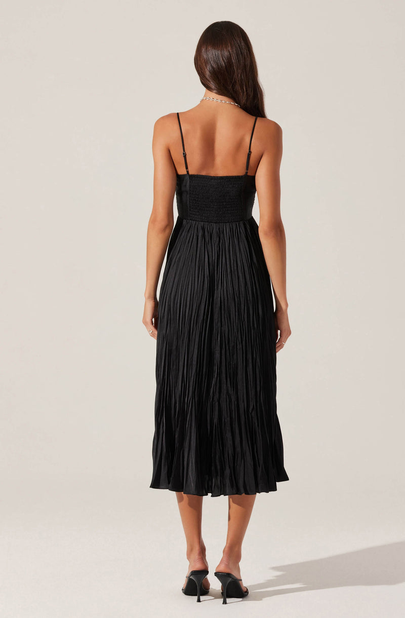 A woman is wearing a black spaghetti strap dress with a fitted bodice and a pleated, mid-length skirt. She is also wearing black high-heeled sandals and a silver necklace.