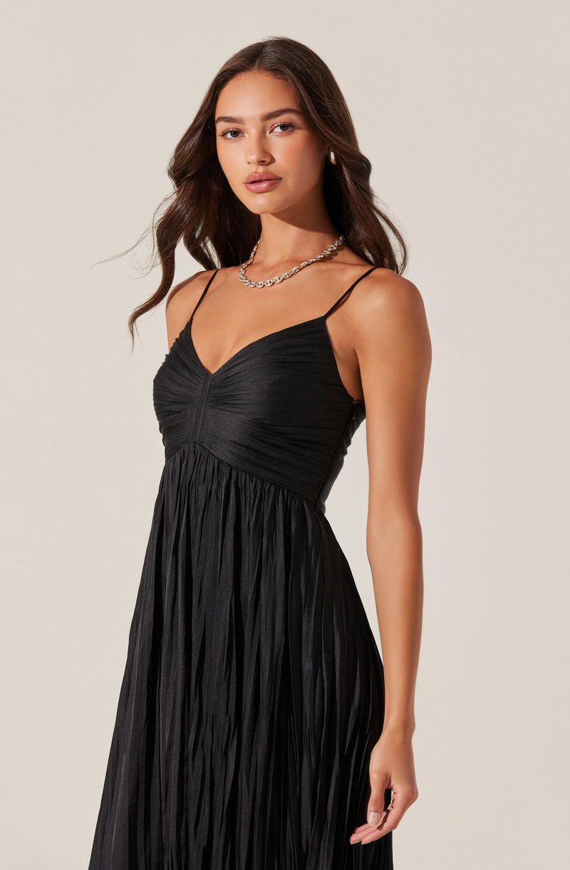A woman is wearing a black, pleated, sleeveless dress with thin spaghetti straps. The dress features a ruched bodice and a flowing skirt. She is accessorized with a silver chain necklace and small earrings.