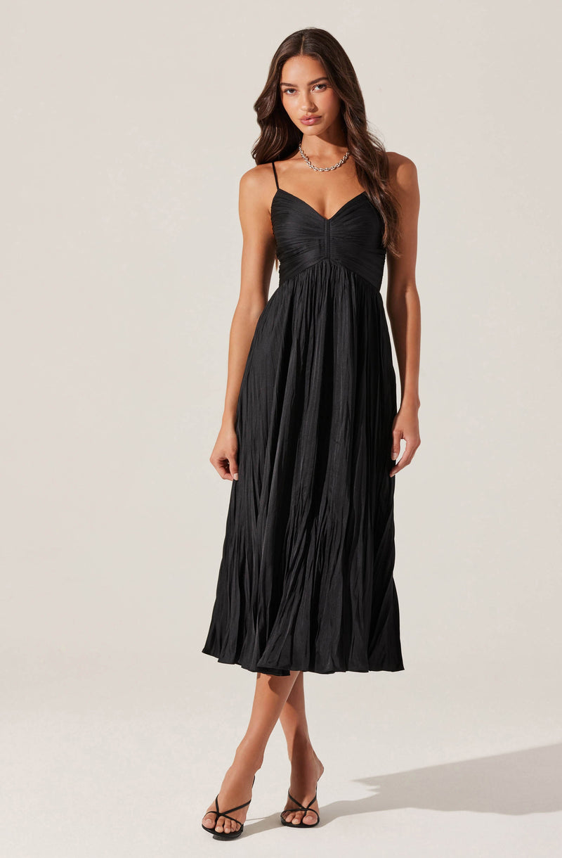 A woman is wearing a black midi dress with thin spaghetti straps. The dress features a ruched bodice and a pleated skirt that falls to mid-calf length. She is accessorizing with a silver chain necklace and wearing black strappy high-heeled sandals.