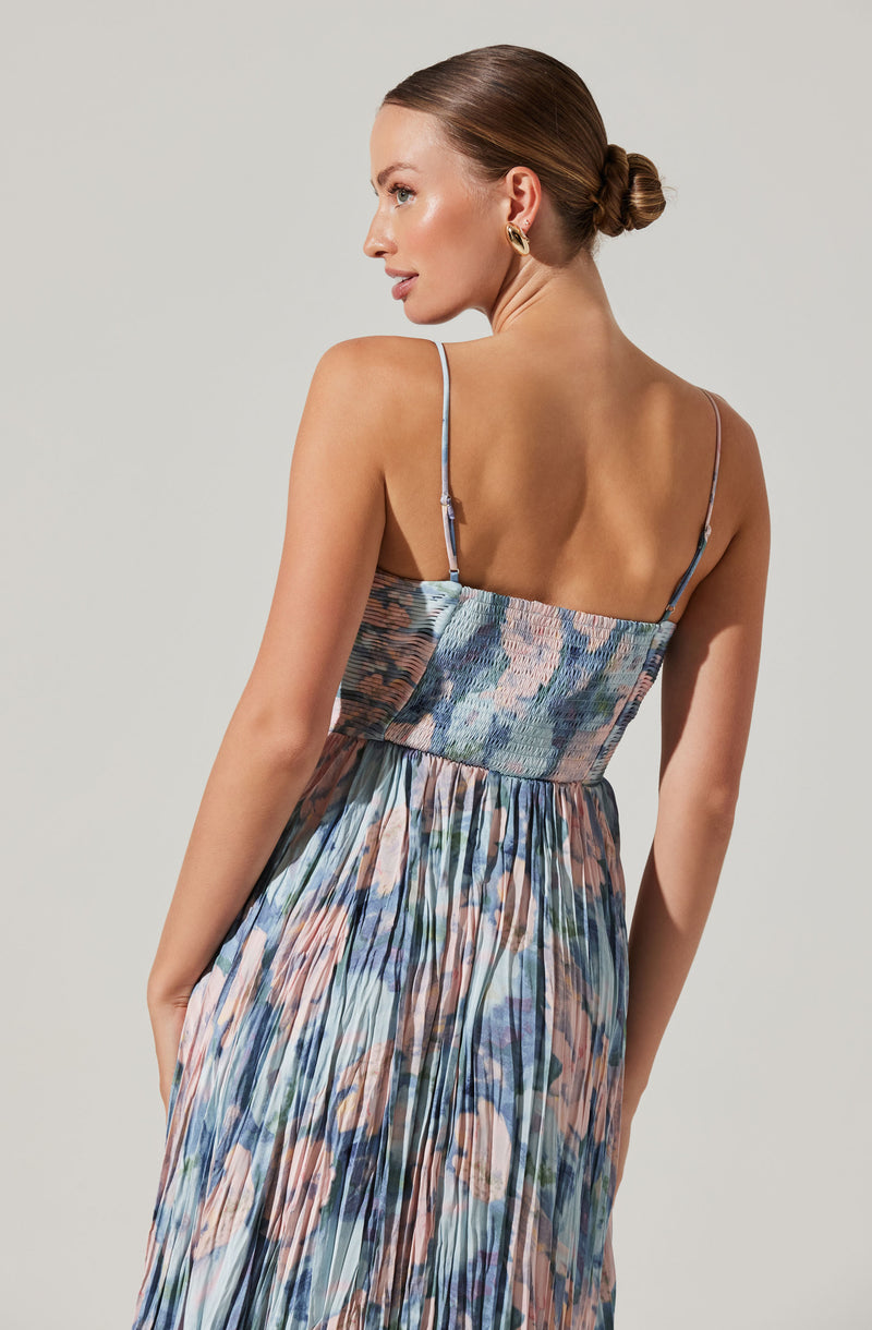 A woman is wearing a sleeveless dress with thin spaghetti straps. The top part is fitted and features a textured, multicolored pattern. The skirt is long, flowy, and has a pleated design with an abstract, multicolored print in shades of blue, pink, and beige.