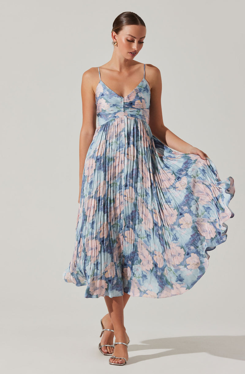 A woman is wearing a sleeveless, V-neck dress with thin spaghetti straps. The dress features a fitted bodice and a flowy, pleated skirt. The fabric has an abstract floral pattern in shades of light pink, blue, and green. She is also wearing silver strappy high-heeled sandals.