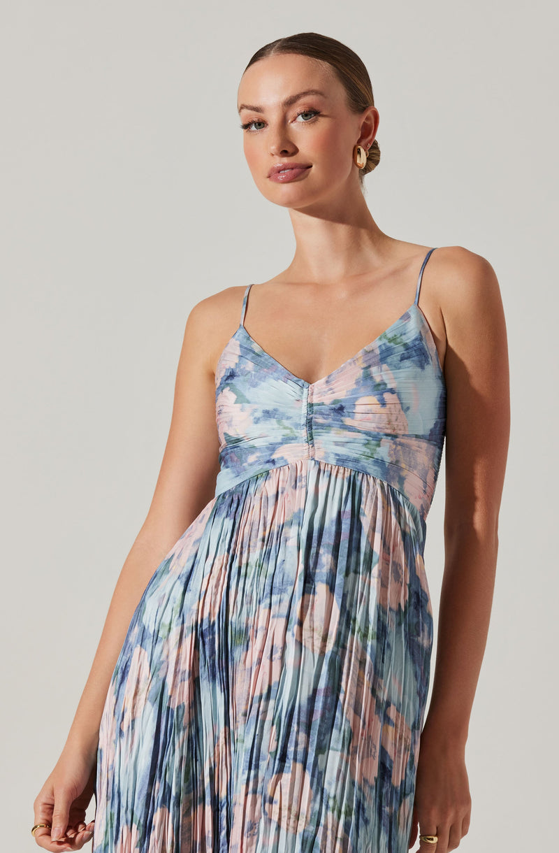 A woman is wearing a sleeveless dress with thin spaghetti straps. The dress features a printed pattern that resembles a watercolor or abstract floral design, with a mix of soft pastel colors including blue, pink, and green. The bodice is fitted with a V-neckline and a subtle front seam, while the skirt is long and flowing with pleats. The overall style of the dress is light and summery.