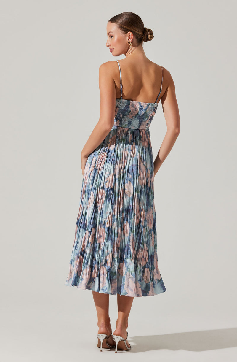 The woman is wearing a sleeveless, ankle-length dress with thin spaghetti straps. The dress features a fitted, smocked bodice and a flowy skirt with a pleated design. The fabric has a multicolored abstract floral pattern with shades of blue, pink, and beige. She is also wearing high-heeled shoes.