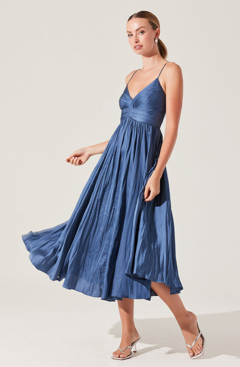 A woman is wearing a sleeveless, pleated, satin dress in a shade of deep blue. The dress features a fitted bodice with a gathered detail at the bust and thin spaghetti straps. The skirt is full and flowing, with a high-low hemline that reveals her legs and her open-toe, strappy heels.