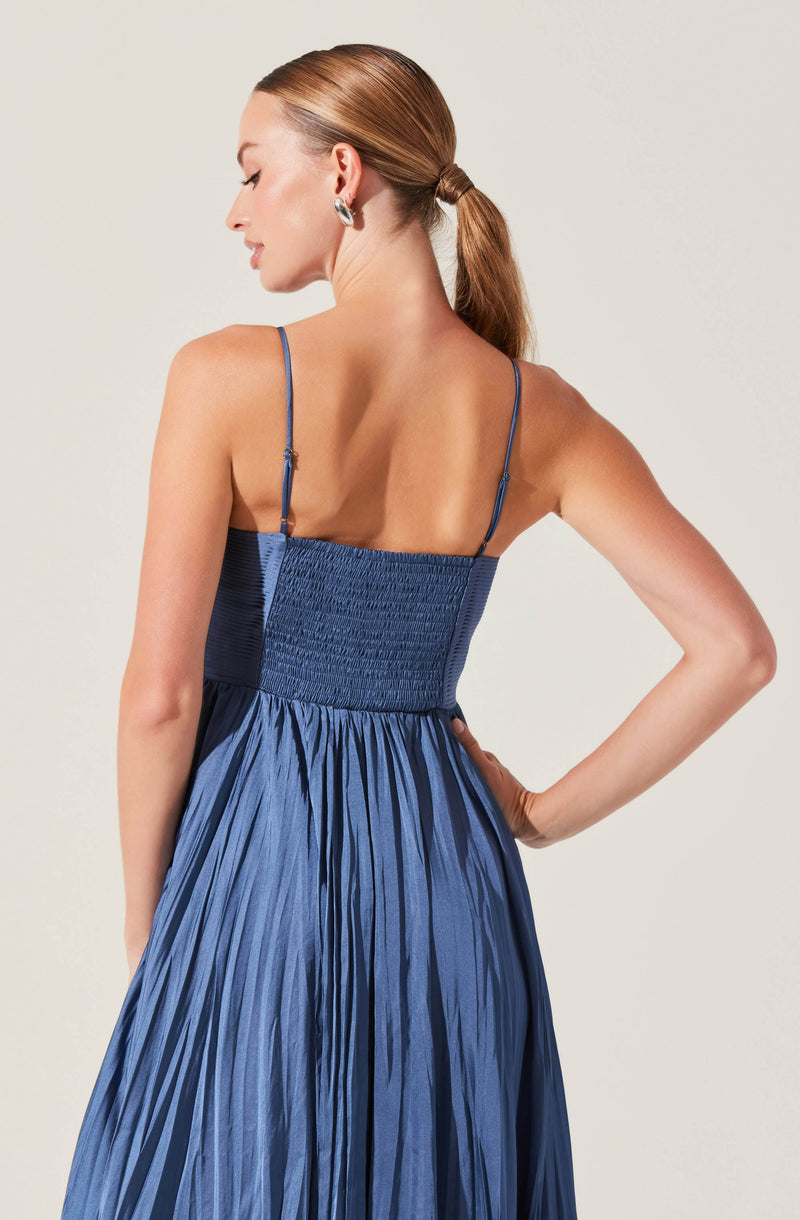 A woman is wearing a sleeveless blue dress with thin, adjustable spaghetti straps. The bodice features smocking with a textured, gathered design, and the skirt is made of shiny, pleated fabric that flows downward.