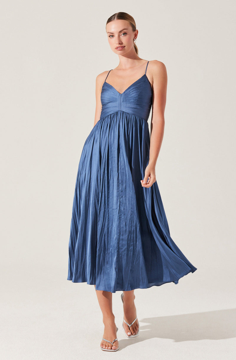 A woman is wearing a sleeveless, deep blue dress with thin spaghetti straps. The dress features a softly ruched bodice and a full, pleated skirt that falls to mid-calf length. She is also wearing a pair of clear, high-heeled sandals.