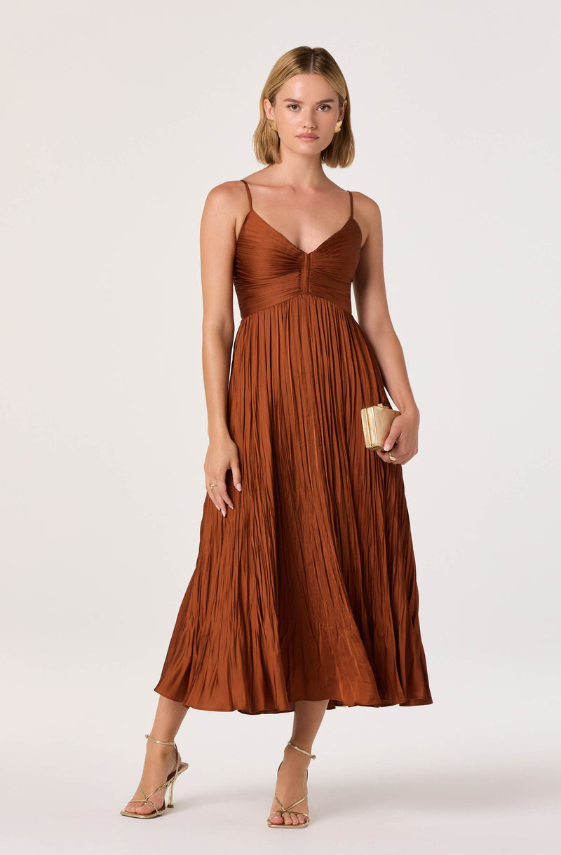A woman is wearing a sleeveless, brown pleated maxi dress with thin spaghetti straps and a fitted bodice. She is holding a small, rectangular clutch purse in her right hand and is wearing high-heeled sandals.