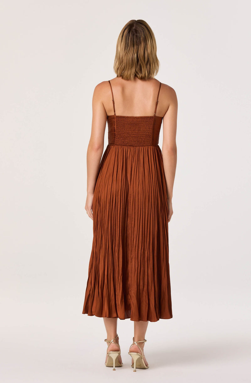 The dress features thin spaghetti straps and is fitted at the waist with a smocked bodice that appears textured. The skirt is long and flowing, with a crinkled, pleated design, and reaches mid-calf length. The fabric is a rich, warm brown color. The model is wearing nude-colored, high-heeled sandals with ankle straps.