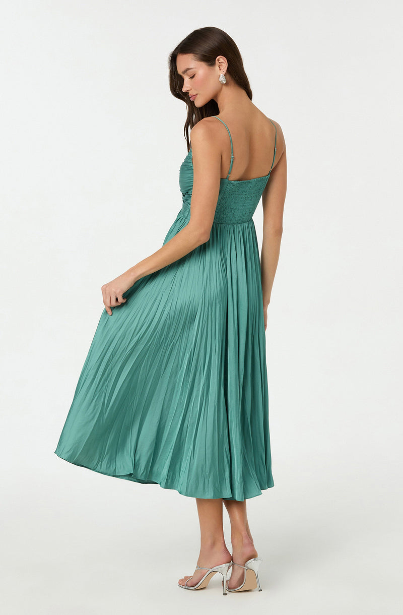 A woman is wearing a sleeveless, teal-colored dress with thin spaghetti straps, a gathered smocked bodice, and a flowing, pleated skirt that reaches mid-calf. She is also wearing high-heeled, open-toe shoes and statement earrings.