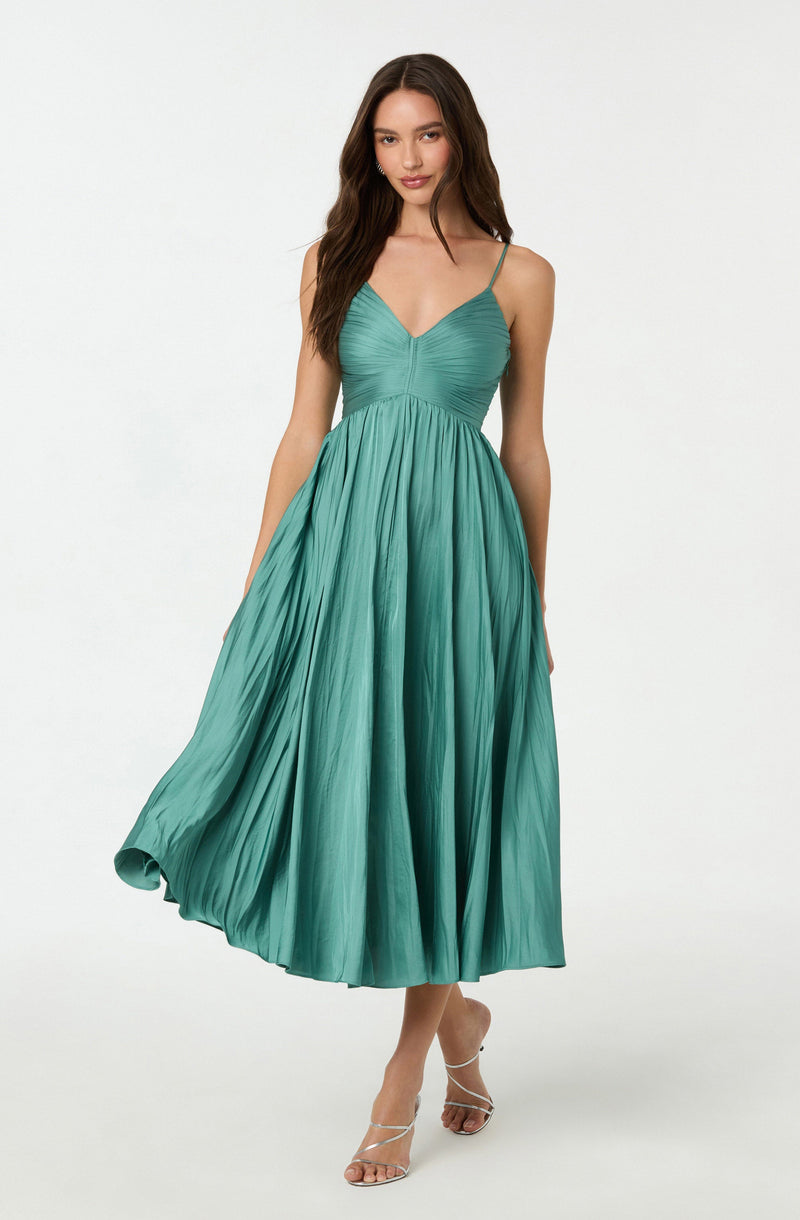A woman is wearing a sleeveless dress with thin spaghetti straps. The dress has a fitted bodice with a gathered detail at the bust. The skirt is long, flowing, and pleated, extending to her ankles with a slight flare. The fabric is smooth and shiny, and the color of the dress is a vibrant shade of teal.