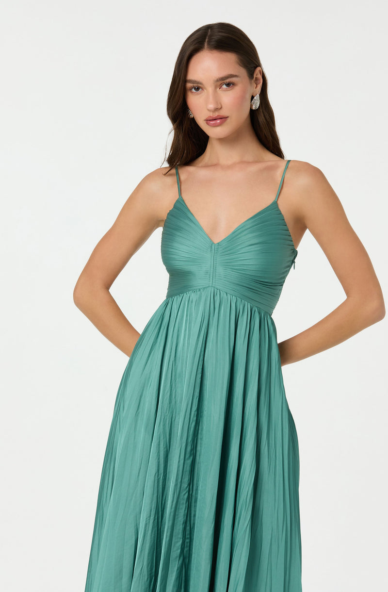 A woman is wearing a sleeveless, teal-colored dress with thin spaghetti straps. The dress features a fitted, ruched bodice with a V-neckline, and a flowing, pleated skirt that extends to the floor. She has long, dark hair and is accessorized with large, round earrings.