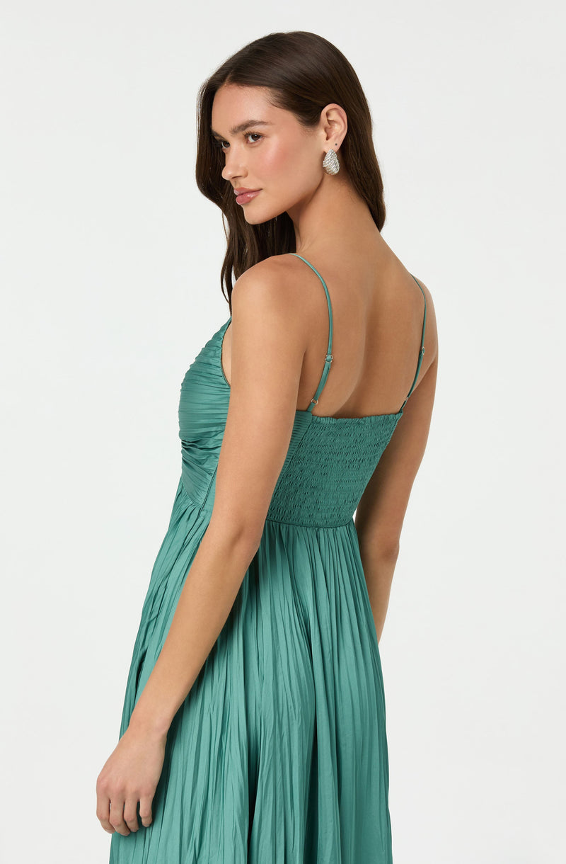 A woman wearing a teal, pleated, sleeveless dress with thin spaghetti straps. The dress has a fitted, textured bodice and flows into a flowing skirt. She is accessorized with large, teardrop-shaped earrings.