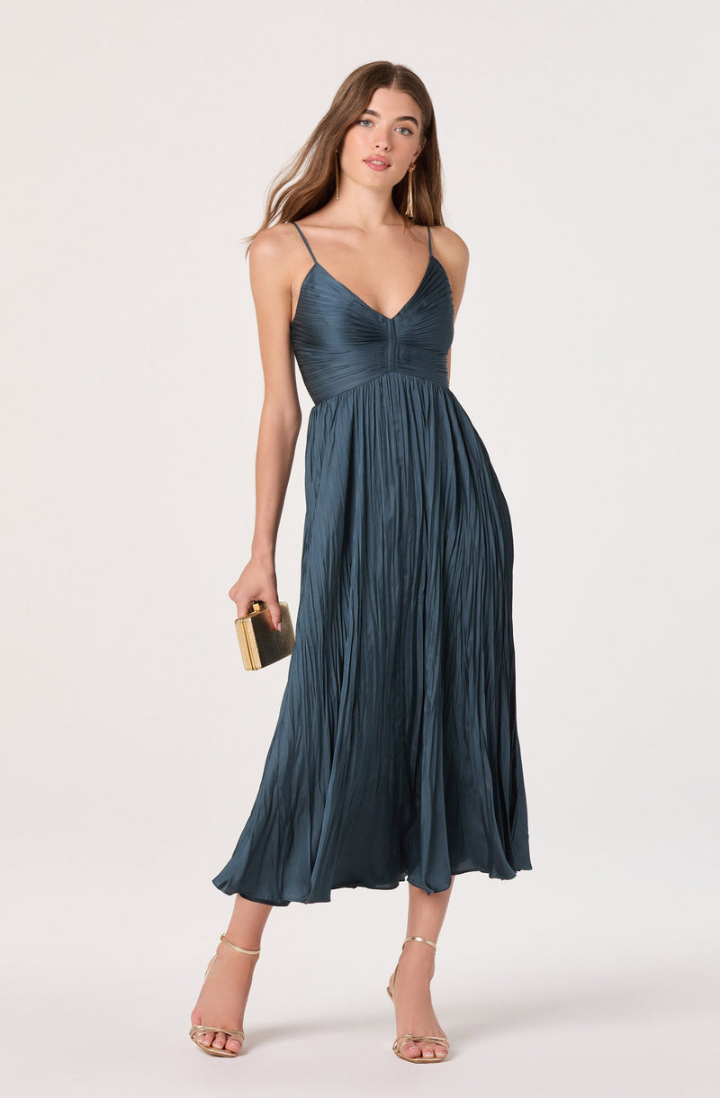 A woman is wearing a dark blue, pleated, sleeveless dress with thin spaghetti straps. The dress has a fitted bodice with a V-neckline and flows down to a midi length. She is holding a small, rectangular clutch and wearing strappy high-heeled sandals.