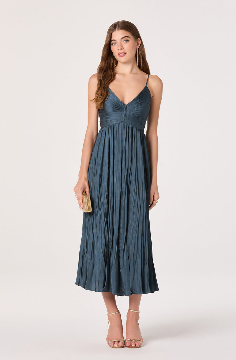 A woman is wearing a long, silky, pleated dress in a deep blue color. The dress has a fitted bodice with thin spaghetti straps and a V-neckline. She is holding a small, gold clutch purse in her right hand and is wearing open-toe high heel sandals.