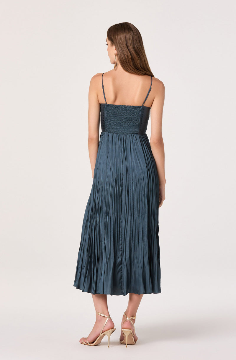 A woman is wearing a dark blue, pleated, sleeveless dress with thin spaghetti straps. The dress has a fitted, smocked bodice and flows down to a midi length. She is also wearing beige high-heeled sandals with thin ankle straps.