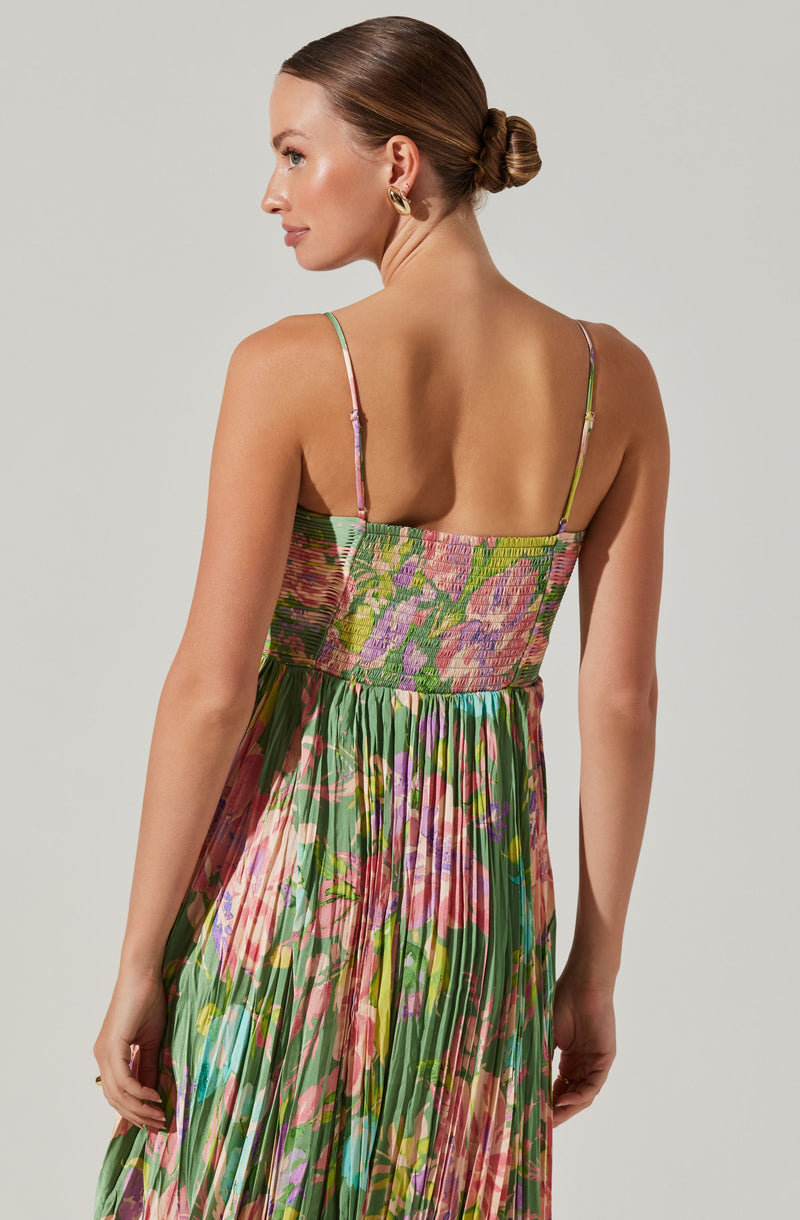 A woman is wearing a sleeveless, ankle-length dress with thin spaghetti straps. The dress features a colorful, floral pattern with shades of pink, purple, green, and hints of blue. The bodice is smocked, providing a fitted look, while the skirt is pleated for a flowing appearance.