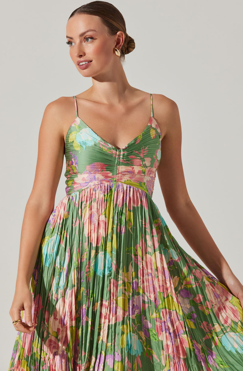 A woman wearing a sleeveless, floral dress with spaghetti straps. The dress has a fitted bodice with a ruched front zipper detail and a full, pleated skirt that features a colorful, vibrant floral pattern with shades of pink, green, purple, blue, and yellow.