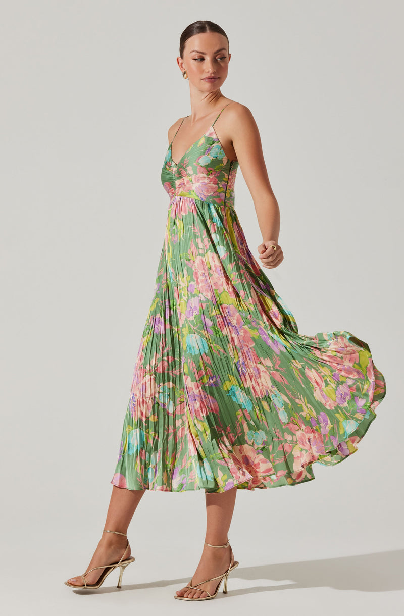 A woman is wearing a sleeveless, V-neck dress with thin spaghetti straps. The dress features a colorful, abstract floral print with shades of pink, green, purple, and yellow. It has a fitted bodice and a flowy, pleated skirt that reaches mid-calf length. She is also wearing nude high-heeled sandals with ankle straps.