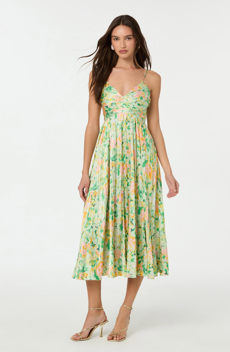 A woman is wearing a sleeveless, ankle-length dress with thin spaghetti straps. The dress has a fitted bodice and a flowing skirt with a vibrant, multicolored floral pattern in shades of yellow, green, pink, and orange. She is also wearing beige high-heeled sandals.