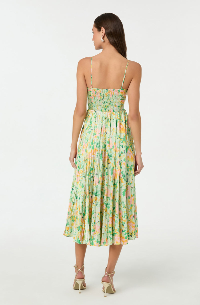 A woman is wearing a sleeveless, ankle-length dress with thin spaghetti straps. The top part of the dress is fitted with a floral pattern, and the skirt features a colorful, abstract floral print with shades of green, pink, and yellow. The dress has a gathered waist and a pleated skirt that falls to mid-calf length. She is also wearing high-heeled sandals with ankle straps.
