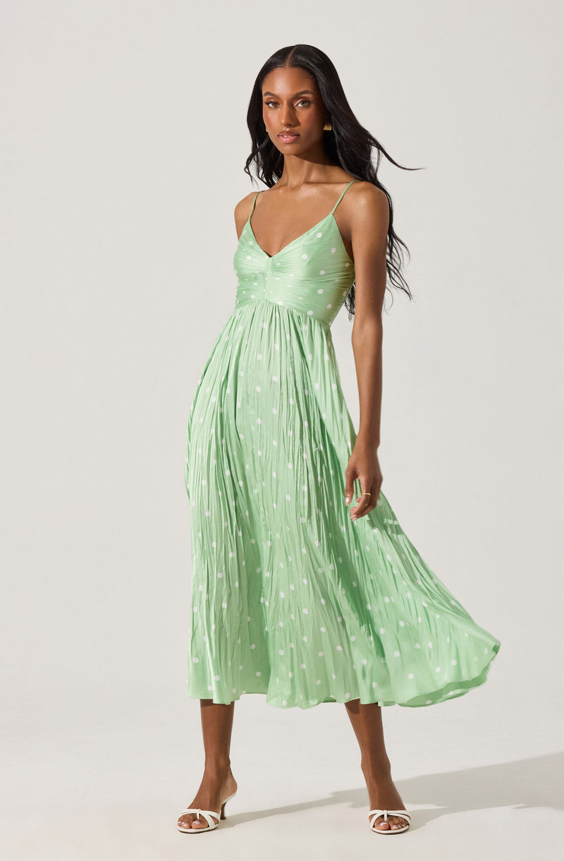 A woman is wearing a sleeveless, light green midi dress with thin spaghetti straps. The dress features a fitted bodice with a V-neckline and a gathered waist, flowing into a pleated skirt. The fabric has a polka dot pattern with white dots scattered throughout. She is also wearing white open-toed high-heeled sandals.