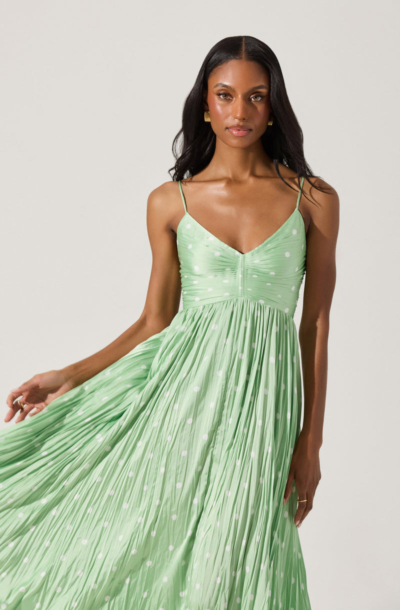 A woman is wearing a light green, pleated maxi dress with thin spaghetti straps. The top of the dress features subtle ruching and a small zippered front detail. The dress has a dotted pattern with white polka dots scattered throughout.