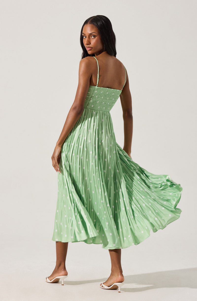 A woman is wearing a light green, pleated, spaghetti-strap dress with a fitted, smocked bodice. The dress has a flowing, midi-length skirt with white polka dot patterns. She is also wearing white high-heeled sandals.