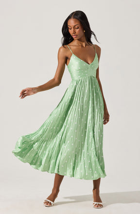 A woman is wearing a light green midi dress with thin spaghetti straps and a V-neckline. The dress features a fitted bodice with a ruched detail at the center and a full, flowing skirt with a pleated texture. The fabric has a subtle polka dot pattern. She is also wearing white open-toe heels.