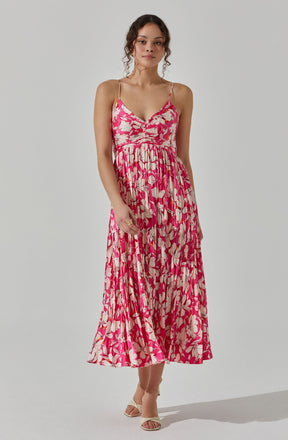 A woman is wearing a sleeveless, pink and beige floral-printed dress with thin spaghetti straps and a V-neckline. The dress has a fitted bodice with a gathered detail at the bust and a flowing, pleated skirt that reaches mid-calf. She is also wearing open-toed, beige high-heeled sandals.