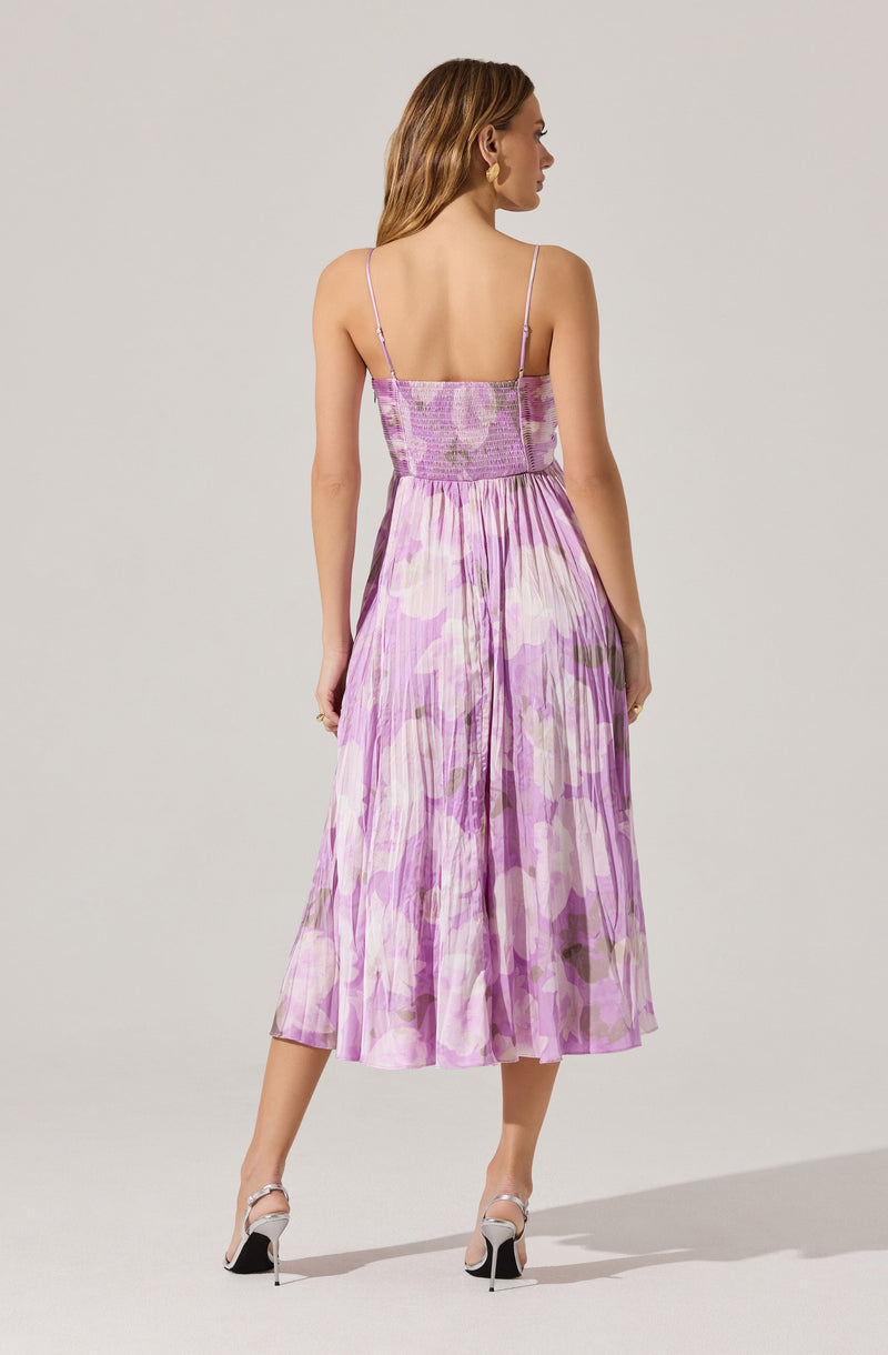 A woman is wearing a sleeveless dress with thin spaghetti straps. The dress features a fitted bodice with shirring at the top and a high-waisted, gathered skirt that falls below the knees. The fabric has a watercolor-like lavender and pink floral pattern with hints of green. She is also wearing high-heeled shoes with ankle straps.