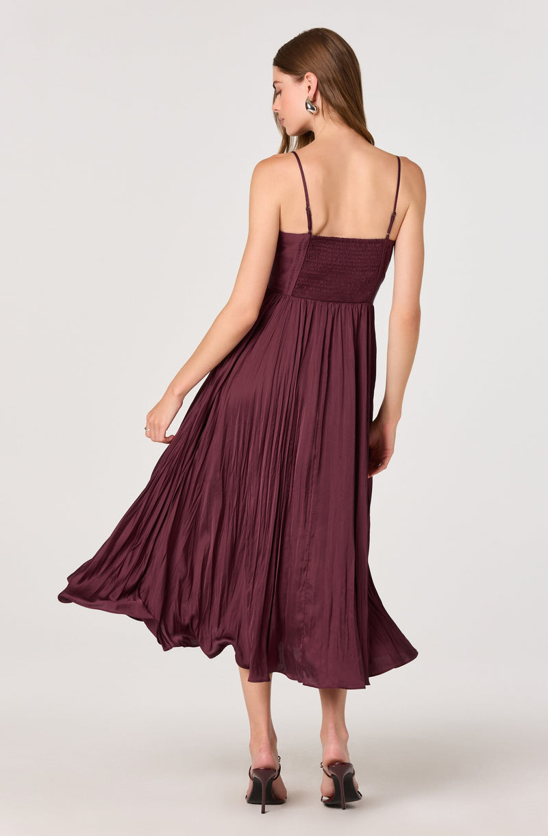 The dress is a sleeveless, ankle-length gown with thin spaghetti straps. It features a fitted bodice with a smocked or ruched detail and a flowing, pleated skirt made of a shiny, satin-like fabric in a deep wine or burgundy color. The dress has a soft, elegant appearance with its gathered skirt creating a voluminous silhouette. The model is also wearing high-heeled sandals with a transparent strap around the toes and a stiletto heel.