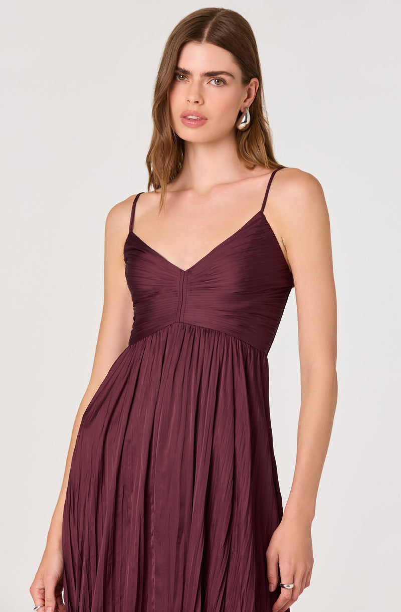 A woman is wearing a sleeveless, deep burgundy dress with thin spaghetti straps. The dress features a fitted, ruched bodice with a V-neckline and a gathered, pleated skirt that flows down.